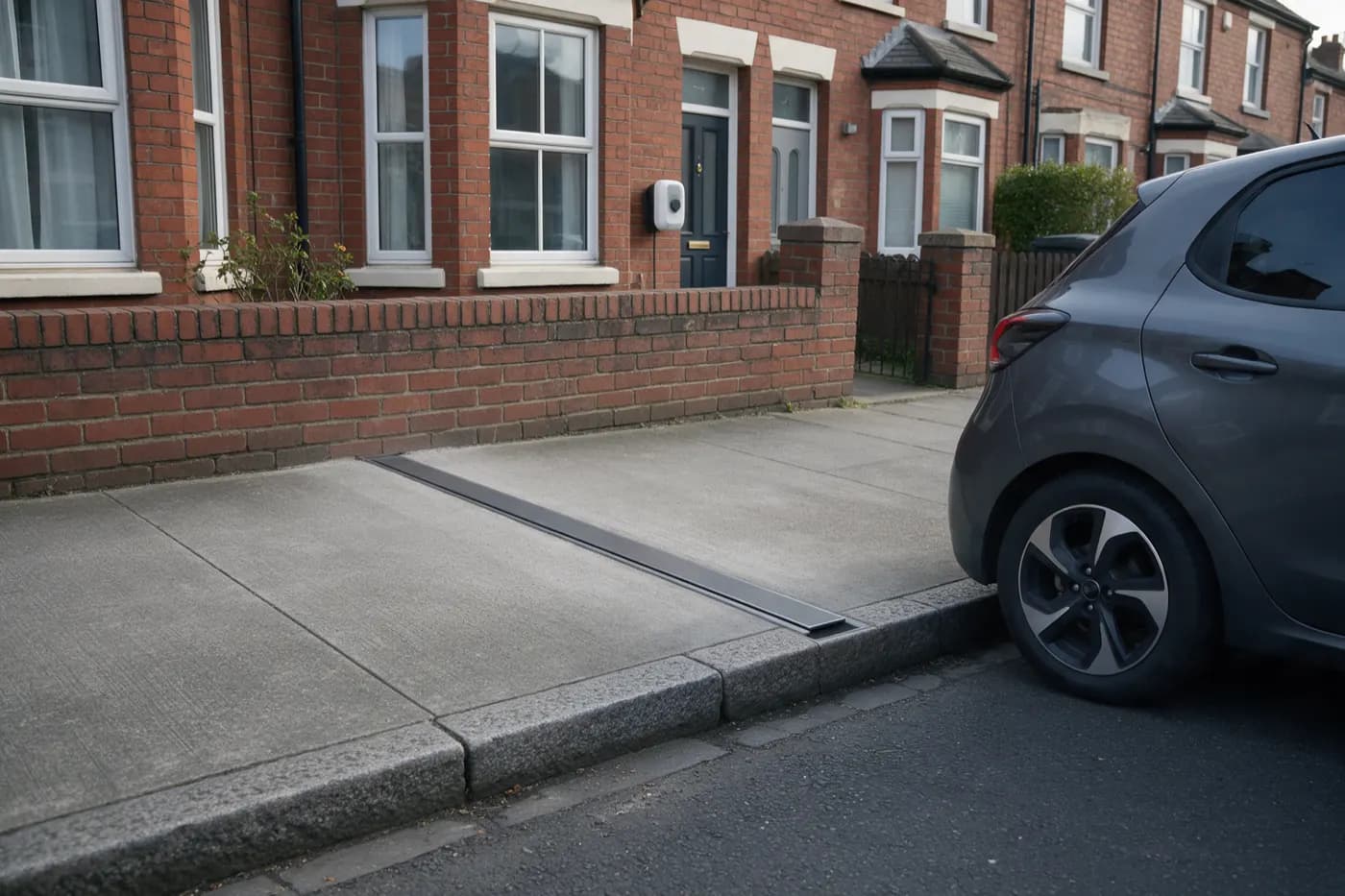 A narrow dark grey cable channel set flush into a concrete pavement, running straight from a terraced house's low brick garden wall to the granite kerb, with a white EV charger on the front wall of the house and a grey electric car parked in the road at the kerb
