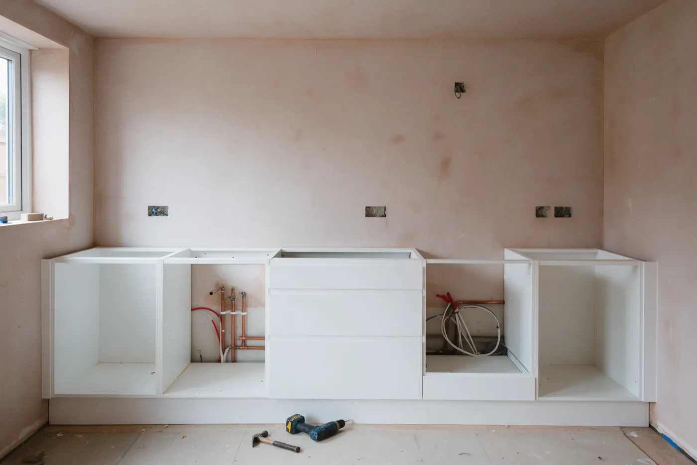 A kitchen part way through fitting: base units in, no worktops yet, pipes and cables waiting