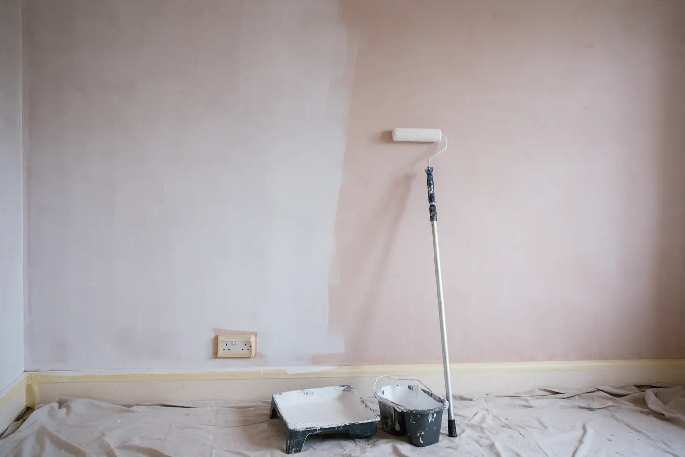 A freshly plastered pale pink wall, the left part covered in a thin, patchy white mist coat and the right part still bare plaster, with a roller on an extension pole leaning on the wall, a roller tray and paint scuttle on a dust sheet, and the socket and skirting board masked with tape