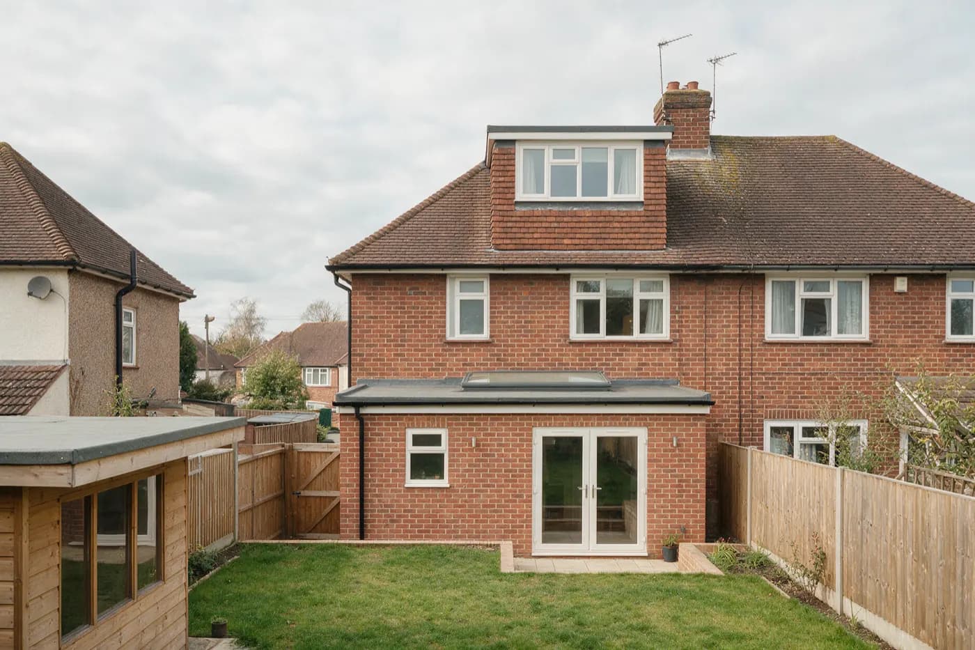 The back of a red-brick semi-detached house with a single-storey flat-roofed rear extension with a rooflight and glazed doors, a tile-hung box dormer on the rear roof, and a timber garden room with a flat roof at the end of the lawn
